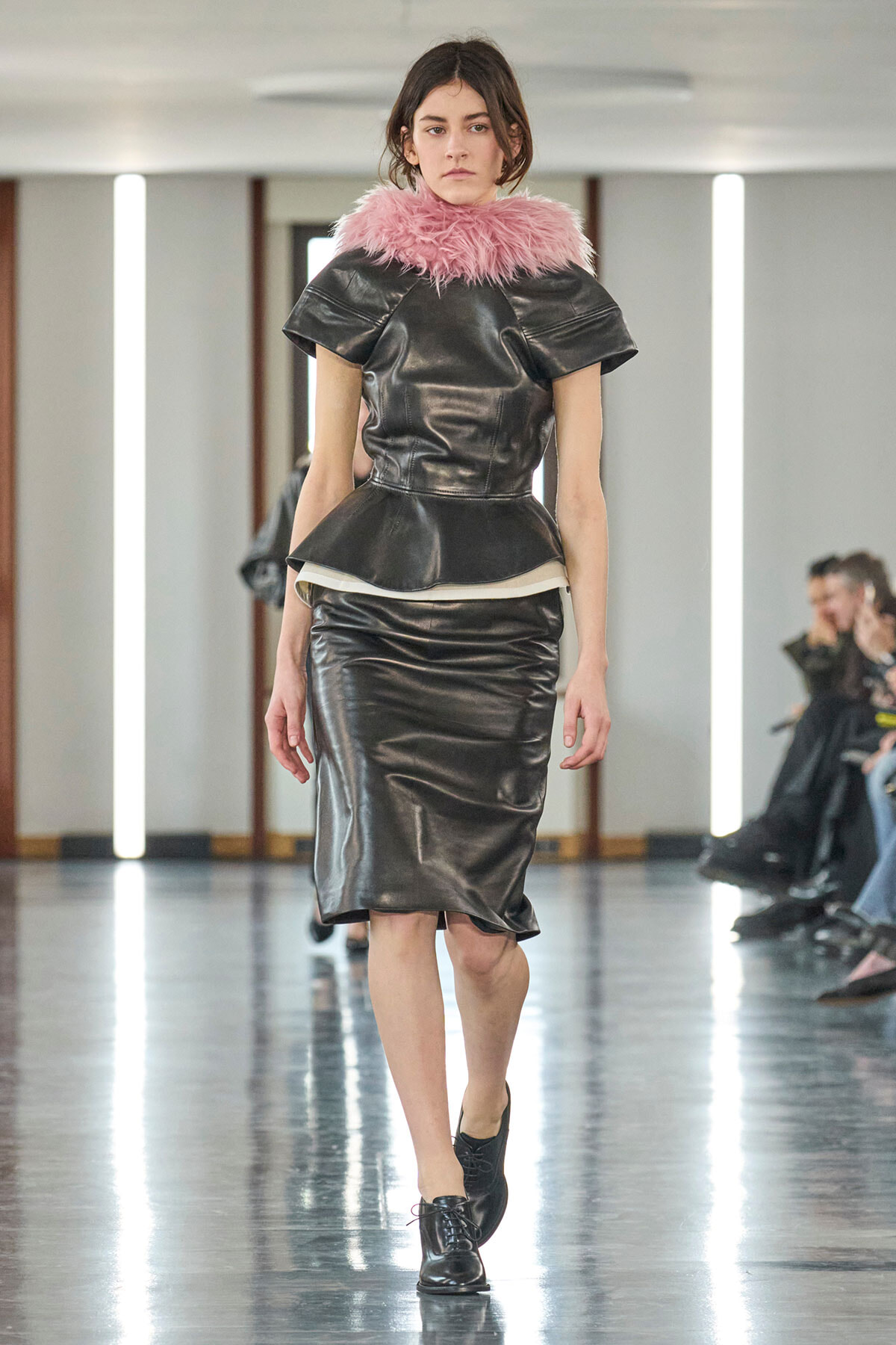 Female model walking runway in a black leather peplum top and matching pencil skirt with a pink feathered collar, black lace-up shoes.