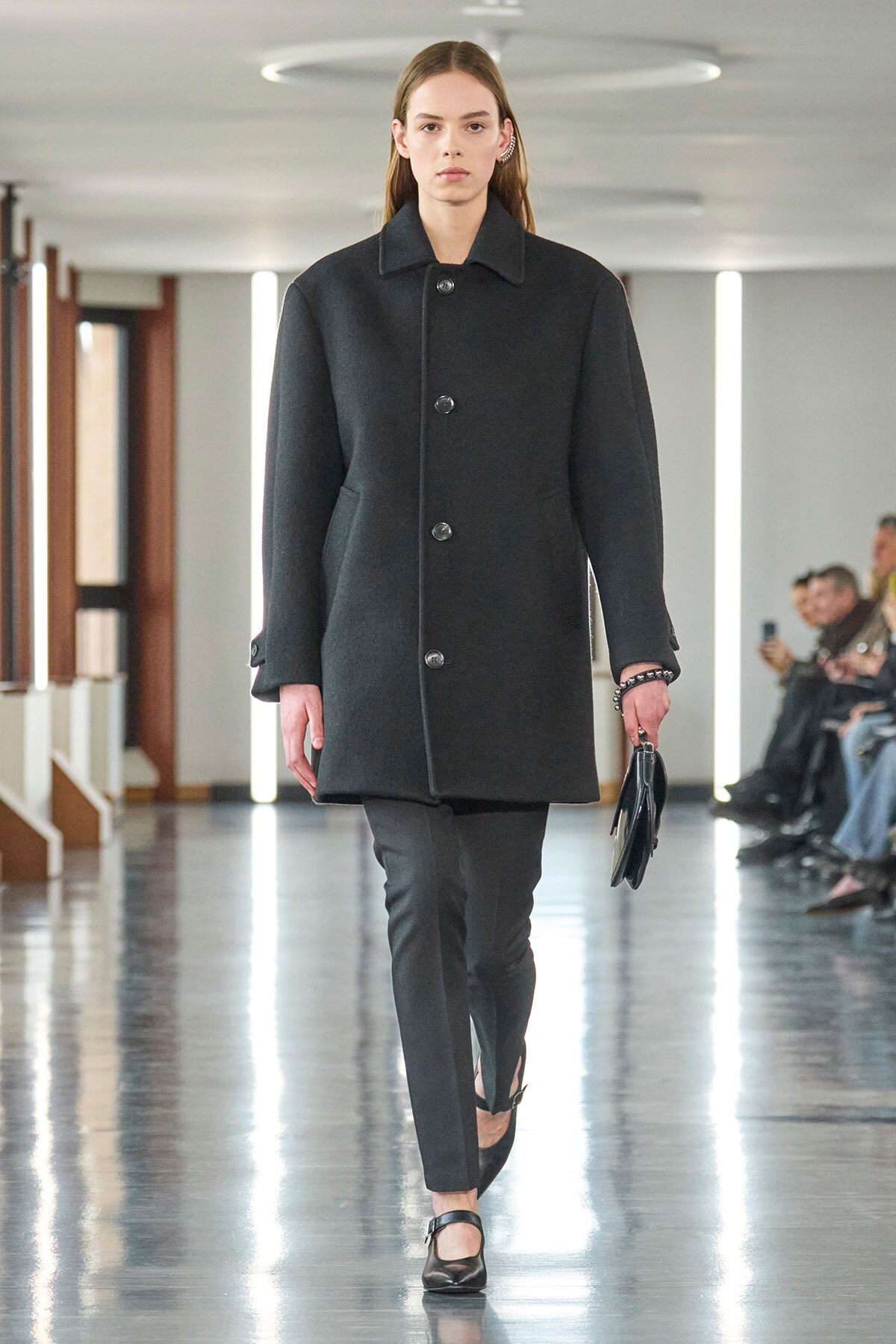 Female model walks a runway in a dark wool coat with large buttons, black pants, and black Mary Jane shoes, holding a small black clutch.
