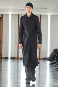 Male model in a knee-length black coat and black beanie walks a fashion runway in an all-black outfit.