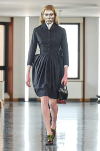 Model walking in a dark, tailored dress with a full skirt, glitter makeup on the face, and a small black handbag in hand.