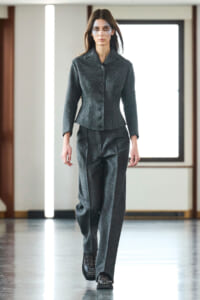 Model wearing a dark grey tailored blazer and wide-leg trousers walking down a runway.