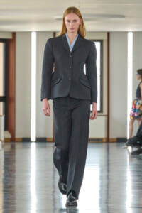 Model walking a runway in a dark gray tailored pantsuit with a light blue shirt, black shoes, in a bright indoor venue.