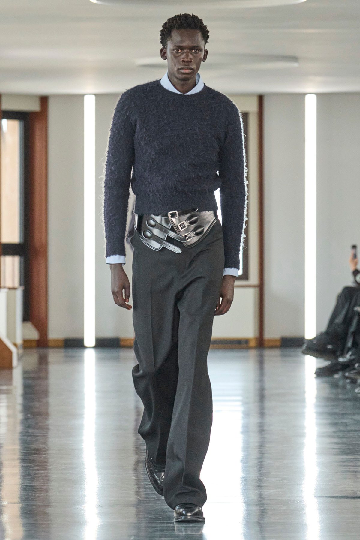 Male model on runway wearing a navy textured sweater over a light collared shirt, with a wide metallic belt, black trousers, and polished black shoes.