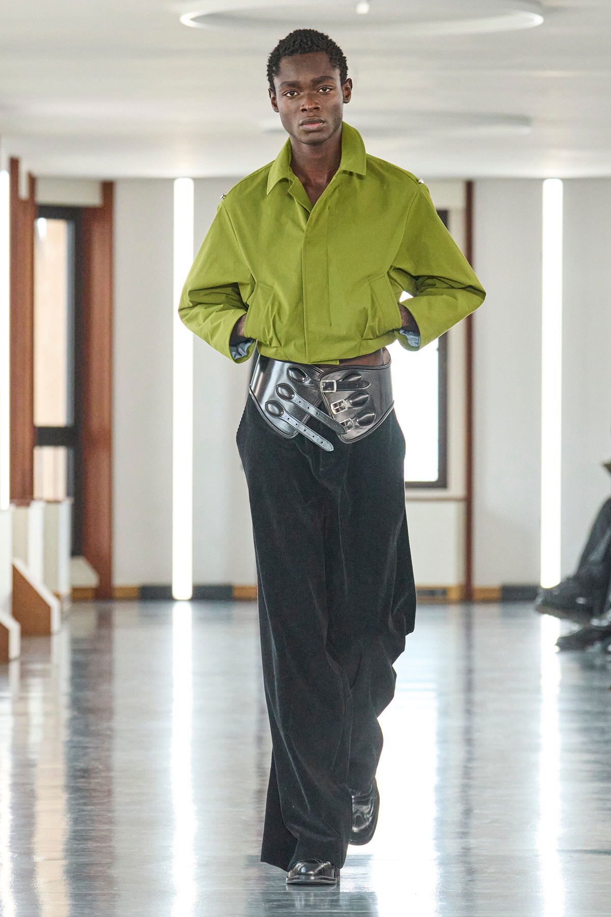 Male model walking a runway in a lime-green oversized jacket, black wide-leg pants, and a metallic waist belt; hands in pockets.