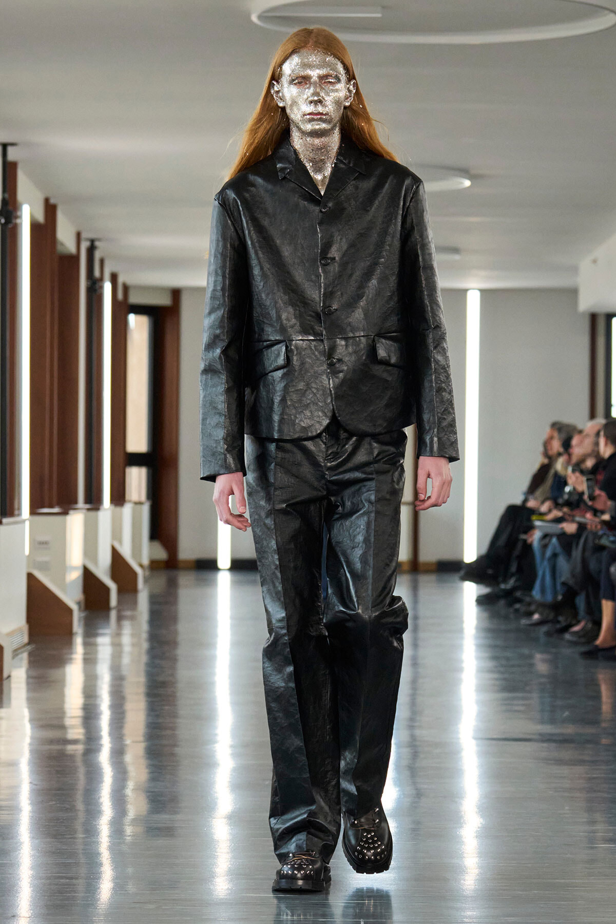 Model walking a runway in a black, glossy leather suit with silver face paint and a neutral expression.