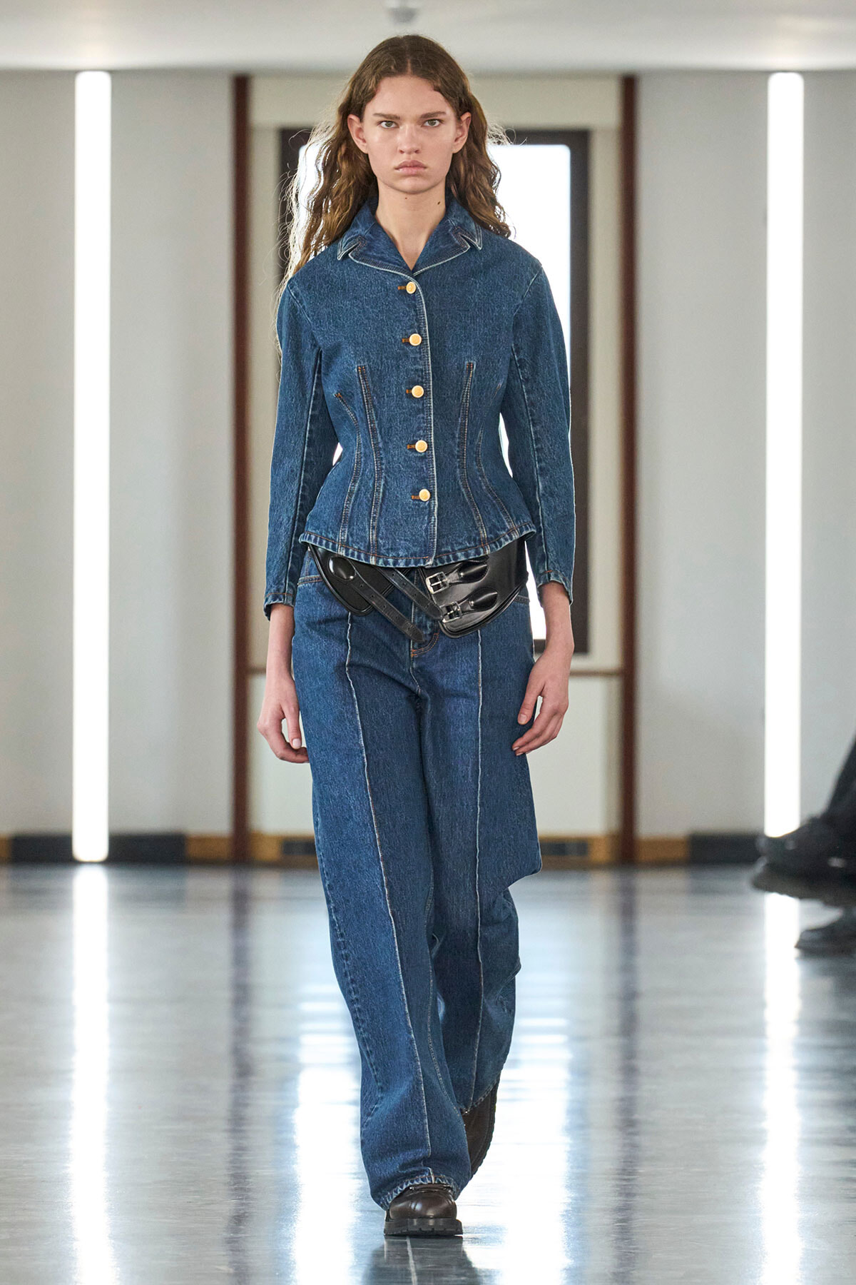 Female model walks a denim-on-denim runway look: cobalt denim jacket with gold buttons and matching wide-leg jeans, cinched with a black leather belt bag, neutral expression in a bright fashion show setting.