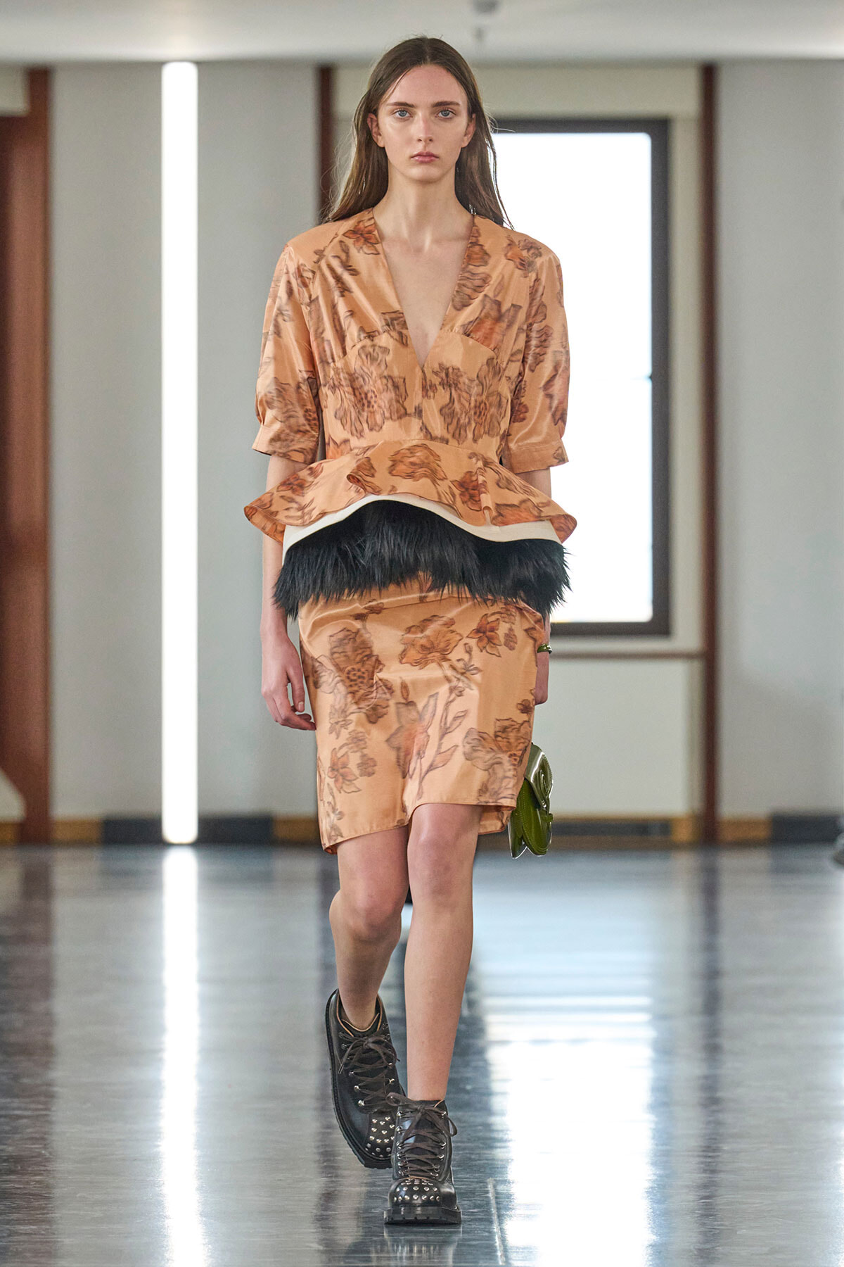 Model walking down the runway in a peach floral dress with a peplum waist and a black feather belt, brown studded boots visible.