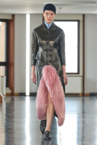 Model walks a runway in a black leather tailored jacket, pink fur skirt, blue shirt, and navy beanie under bright studio lights.