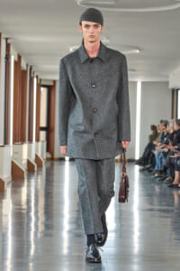 Male model on a runway wearing a grey wool coat and matching trousers, black cap, and carrying a burgundy leather bag while audience watches.