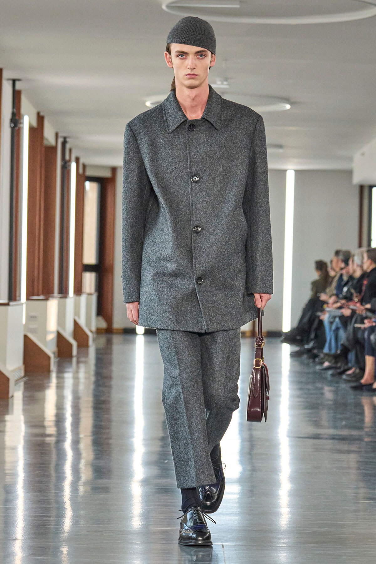 Male model on a runway wearing a grey wool coat and matching trousers, black cap, and carrying a burgundy leather bag while audience watches.