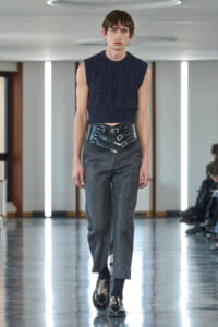 Model walking the runway in a navy sleeveless knit top with a wide black buckled belt over gray trousers.