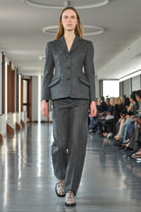 Female model walks a gray tailored pantsuit down a fashion-show runway with seated audience along the sides.