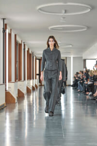Model walking a glossy hallway runway in a charcoal grey tailored suit, with a seated audience along the right taking photos.