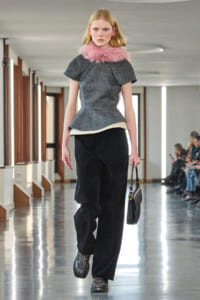 Model walking a runway in a gray textured top with a pink fur collar, black wide-leg pants, studded boots, and a small black handbag.