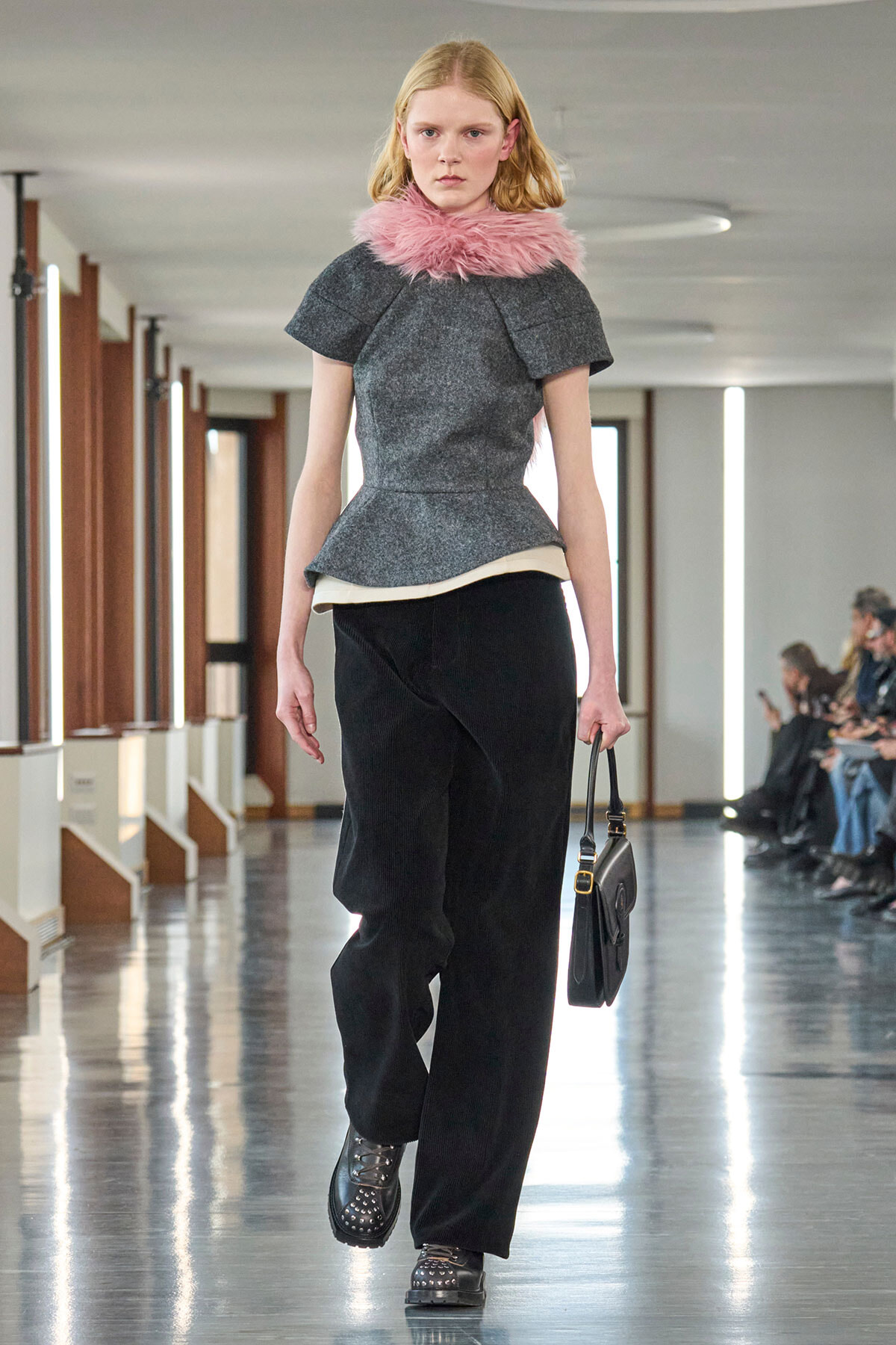 Model walking a runway in a gray textured top with a pink fur collar, black wide-leg pants, studded boots, and a small black handbag.