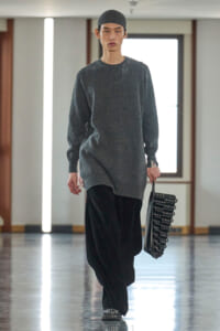 Male model walking a runway in an oversized charcoal gray sweater, black wide-leg pants, and a gray knit cap, carrying a black studded handbag.