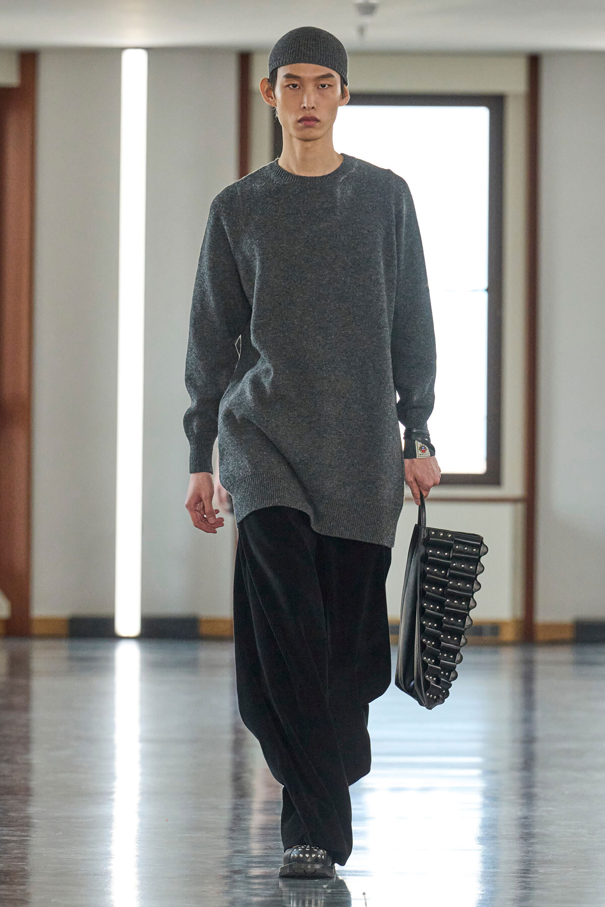 Male model walking a runway in an oversized charcoal gray sweater, black wide-leg pants, and a gray knit cap, carrying a black studded handbag.