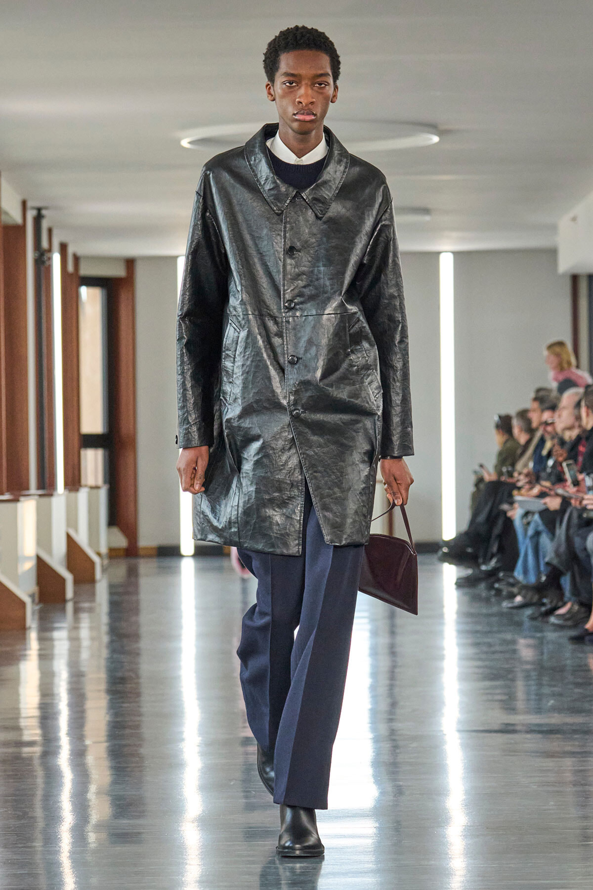 Male model walks a runway in a shiny black oversized coat, navy trousers, and black boots, carrying a burgundy clutch.