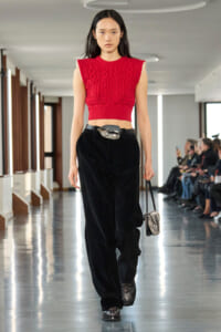 Model walks a fashion runway wearing a red cropped knit top, black velvet wide‑leg pants, a wide black belt with a large buckle, chunky studded boots, and a small snakeskin bag. Audience in the background.]','Model on a runway in a red sleeveless knit crop top and black velvet pants, carrying a small snakeskin purse.