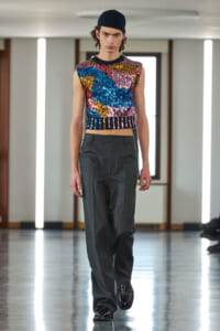 Model walking a runway in a sleeveless multicolor sequin crop top, dark gray wide-leg trousers, and a navy beanie.