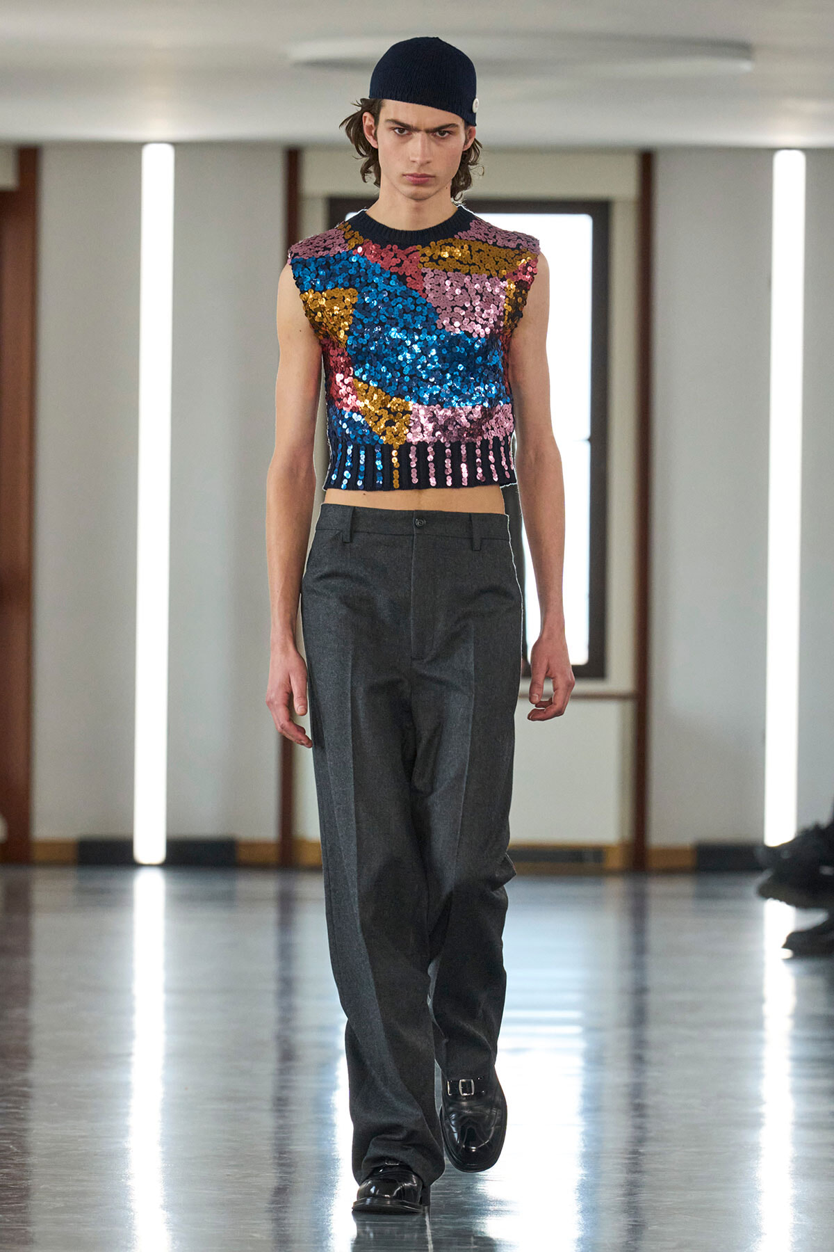 Model walking a runway in a sleeveless multicolor sequin crop top, dark gray wide-leg trousers, and a navy beanie.