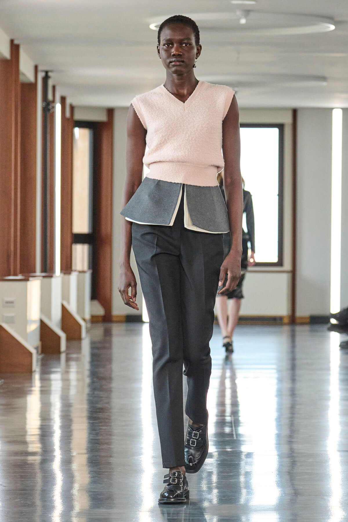 Model walking a runway in a light pink sleeveless knit top with a layered peplum over dark trousers and black buckle shoes in a bright hallway.