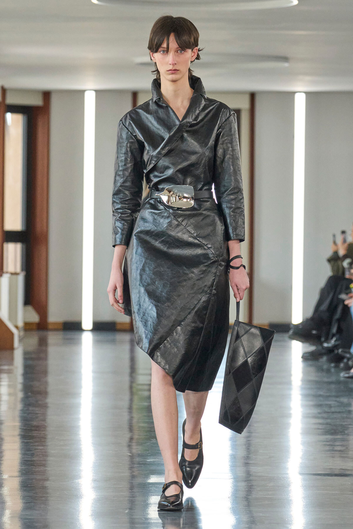 Model on a runway wearing a black metallic wrap dress with a wide silver belt and a black quilted clutch nearby her side.