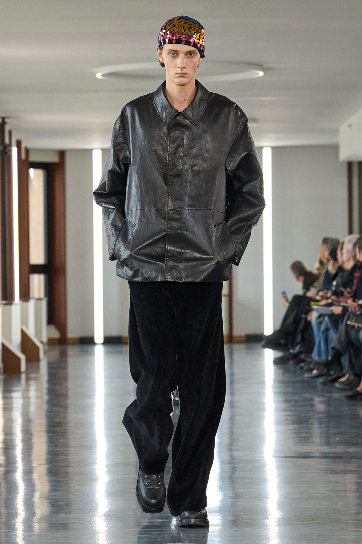 Male model walking a runway in a shiny, oversized black leather jacket, black wide-legged pants, and a colorful sequined cap.