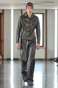 Model walking a runway in a black leather-style matching jacket and wide pants, wearing a colorful sequined headscarf to accessorize.