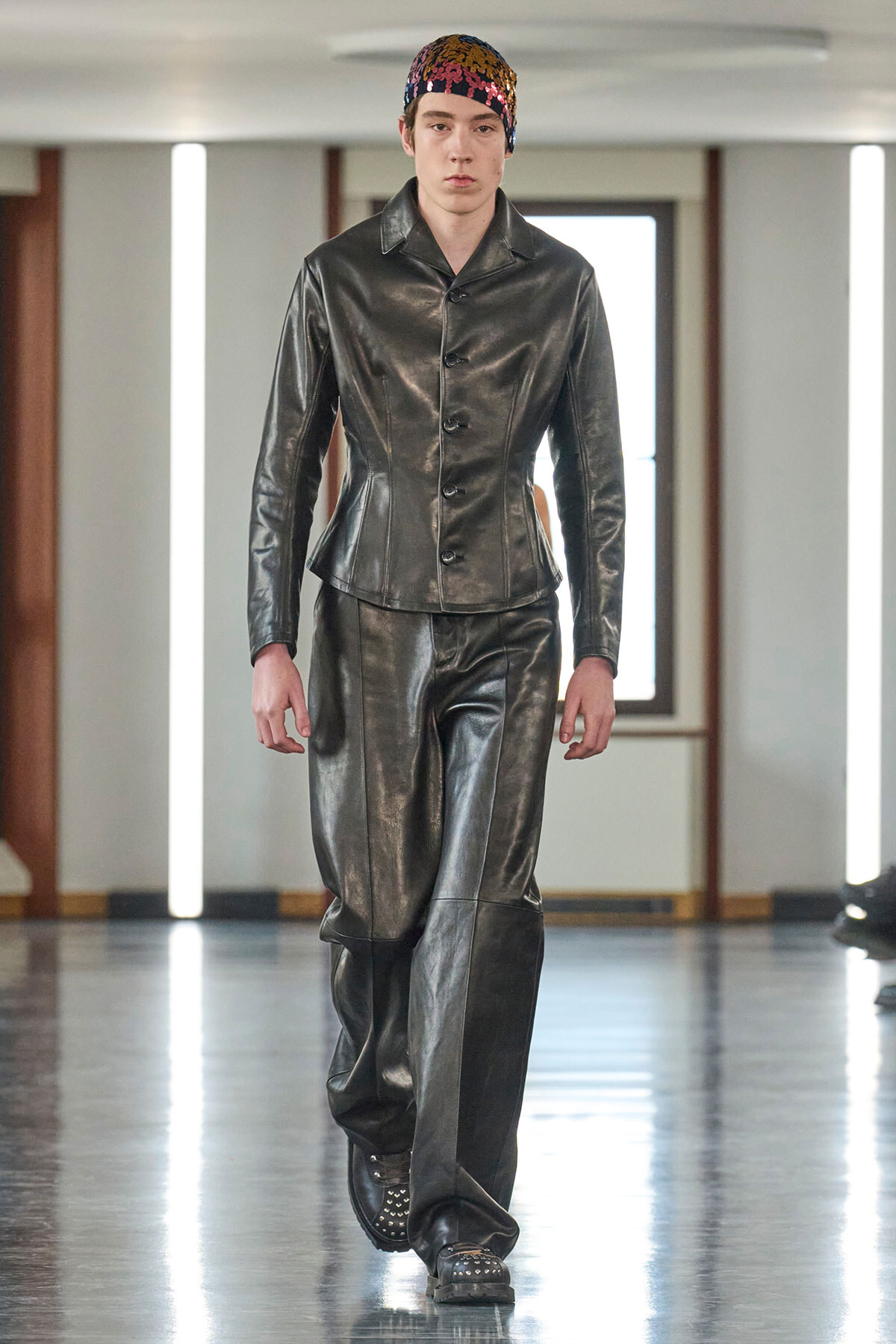 Model walking a runway in a black leather-style matching jacket and wide pants, wearing a colorful sequined headscarf to accessorize.