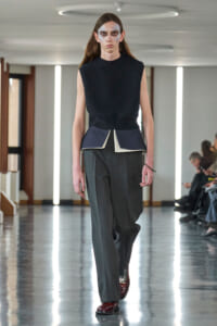 Model walks a runway wearing a black sleeveless top with layered peplum over wide gray trousers and burgundy shoes, with dramatic pale makeup.
