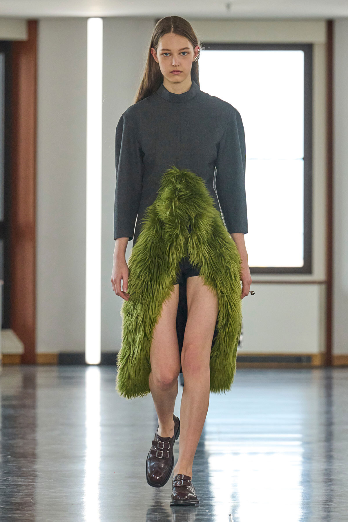 Model walking a runway in a dark high-neck top with a lime-green feathered front garment and brown double-buckle loafers.