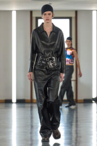Model walking a fashion show in a black leather shirt and pants with a wide sculpted belt and navy beanie.