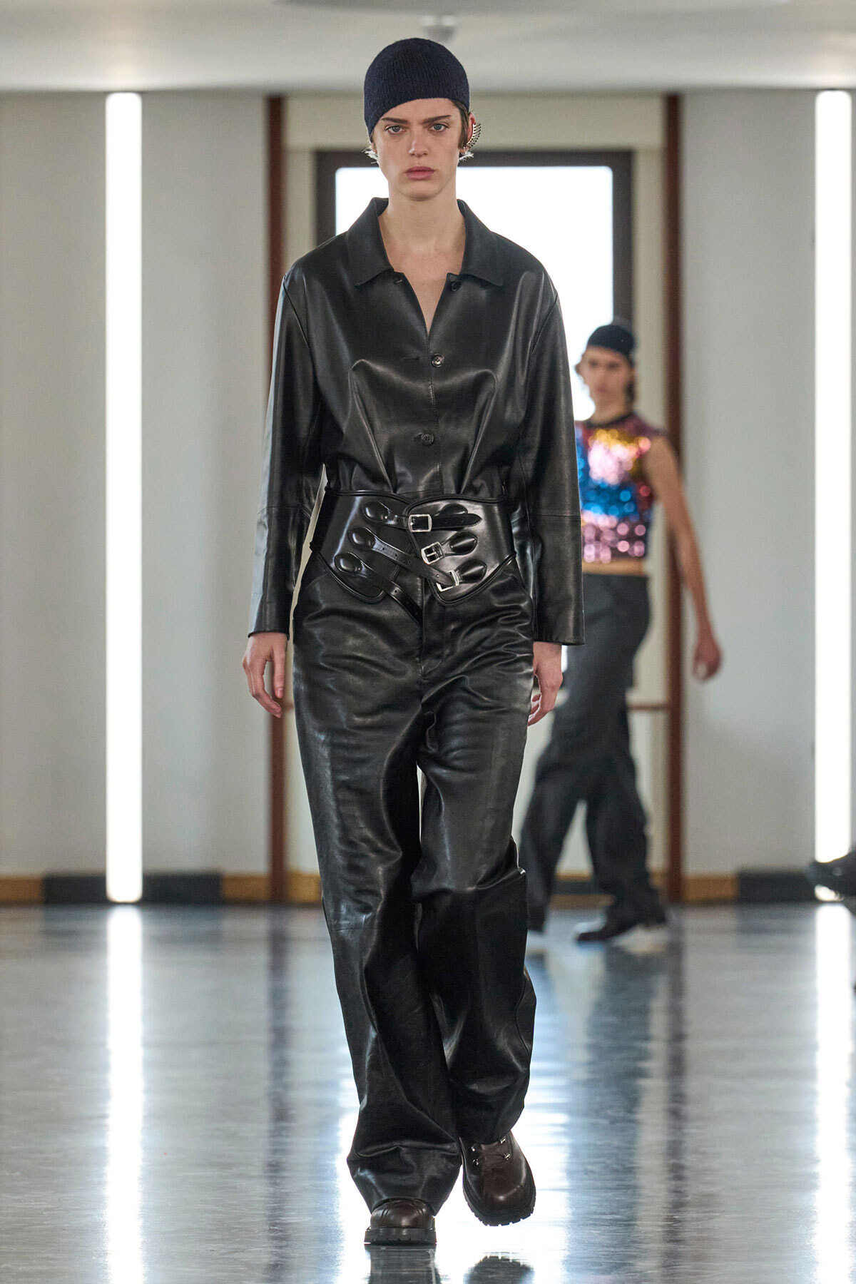 Model walking a fashion show in a black leather shirt and pants with a wide sculpted belt and navy beanie.