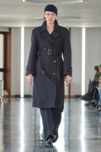 Model walking a fashion runway in a dark double‑breasted trench coat with a waist belt, black beanie, and black boots on a glossy floor.
