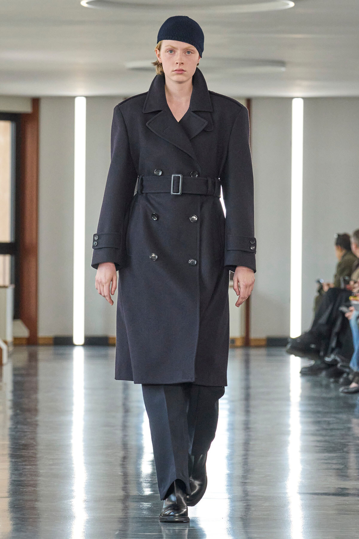 Model walking a fashion runway in a dark double‑breasted trench coat with a waist belt, black beanie, and black boots on a glossy floor.