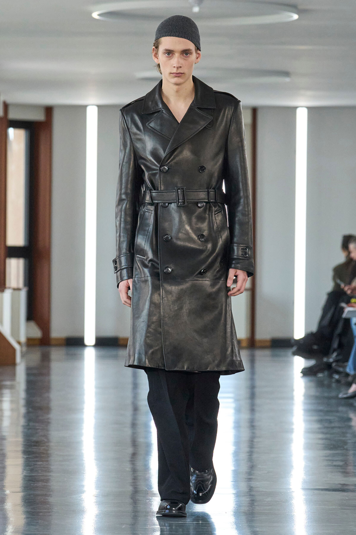 Runway model wearing a black leather trench coat with a belt and dark trousers in a modern showroom setting.