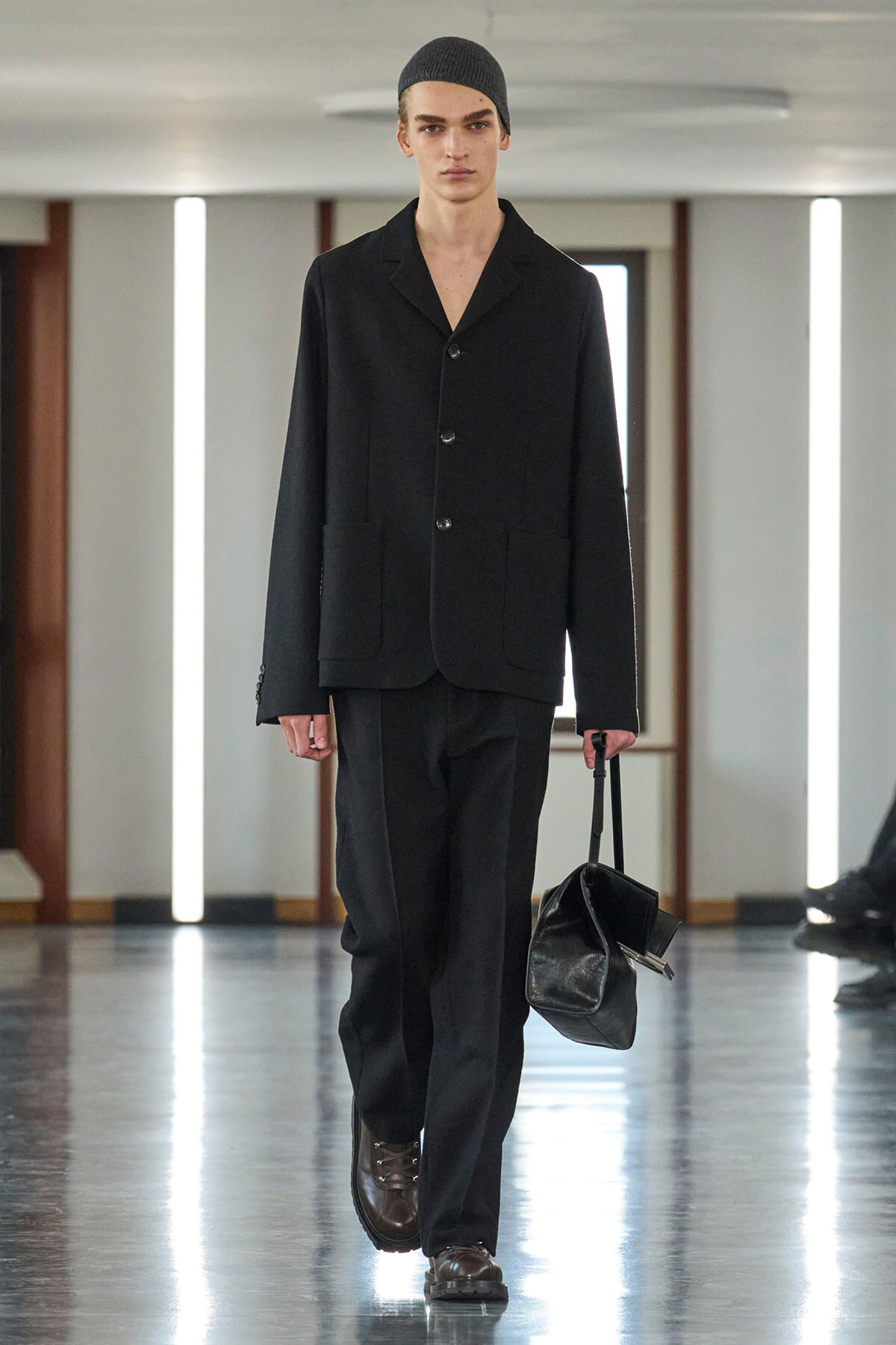 Male model in a black suit and gray beanie walking a runway, carrying a black leather bag.