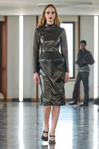 Model in a glossy black leather midi dress with long sleeves and high neck walks the runway in black Mary Jane shoes.