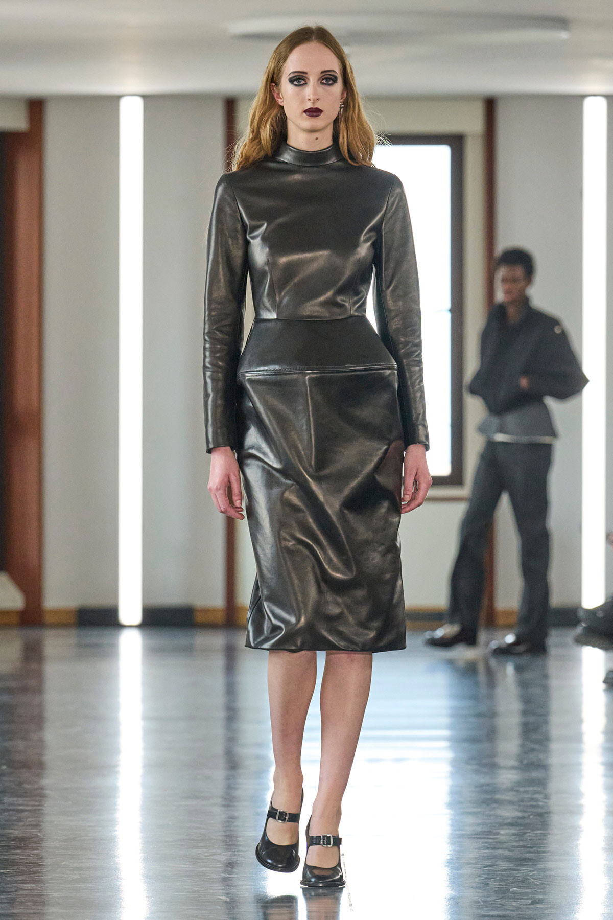 Model in a glossy black leather midi dress with long sleeves and high neck walks the runway in black Mary Jane shoes.