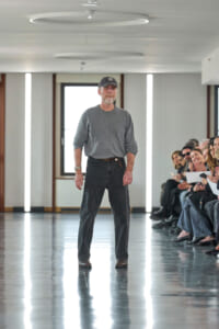 A man in a gray long-sleeve shirt and dark jeans walks down an indoor runway as a seated audience watches on the right.
