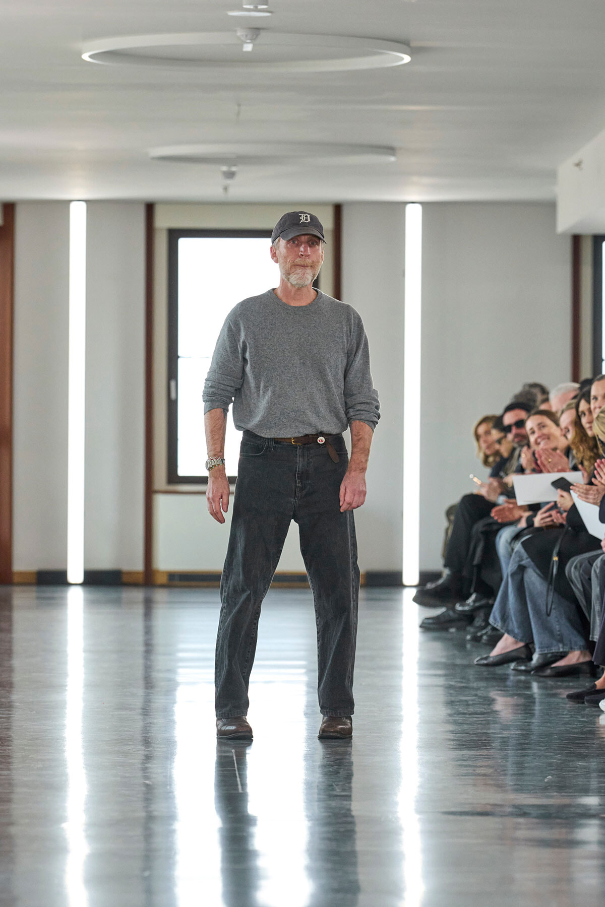 A man in a gray long-sleeve shirt and dark jeans walks down an indoor runway as a seated audience watches on the right.