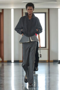 Model walks a fashion runway wearing a loose black jacket layered over a gray peplum and matching trousers, glossy shoes visible.