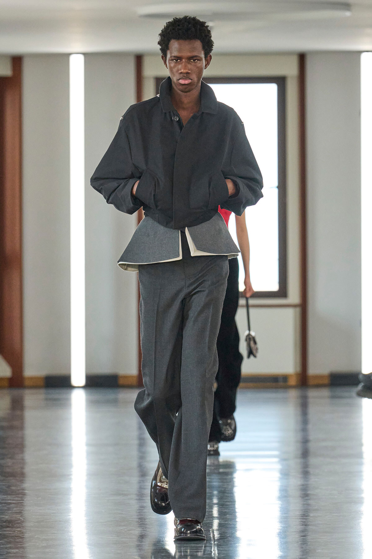 Model walks a fashion runway wearing a loose black jacket layered over a gray peplum and matching trousers, glossy shoes visible.