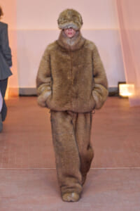 Model on the runway in a bulky brown fur outfit with hood; hands in pockets and face obscured by the fur hood.