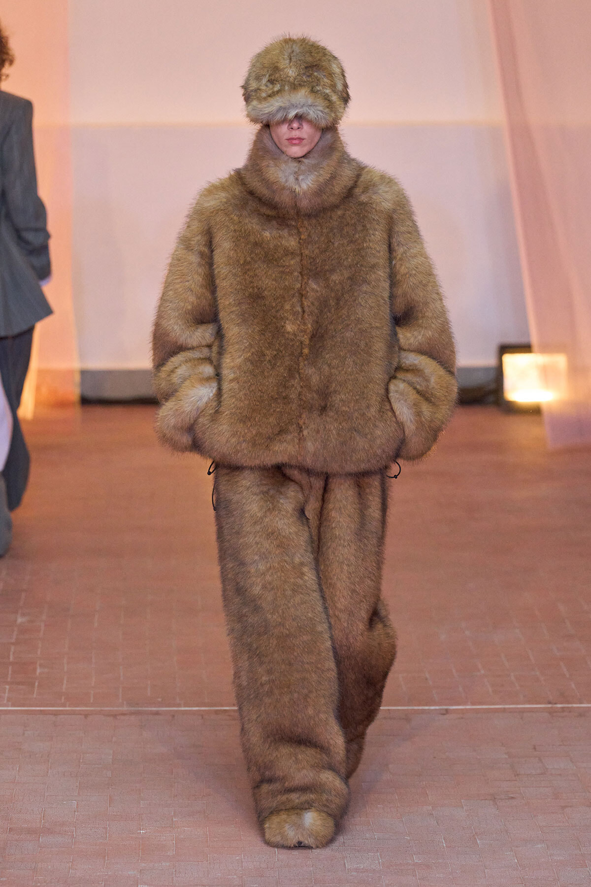 Model on the runway in a bulky brown fur outfit with hood; hands in pockets and face obscured by the fur hood.