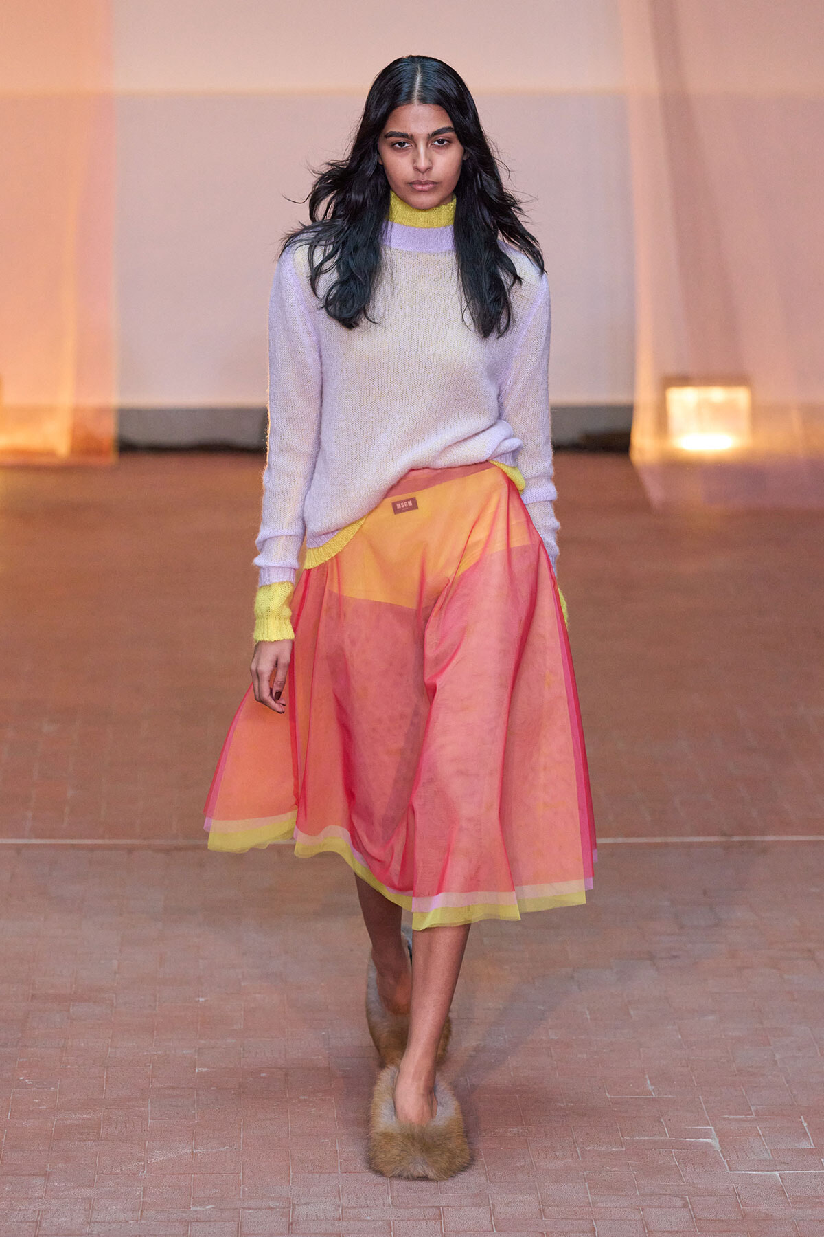 Model walking a runway in a lavender knit sweater with neon yellow trim and a sheer pink-orange midi skirt with yellow hem, wearing fuzzy fur slippers.