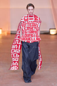 Model walking a runway in a bold red-and-white geometric knit poncho with a cape, paired with dark denim jeans.