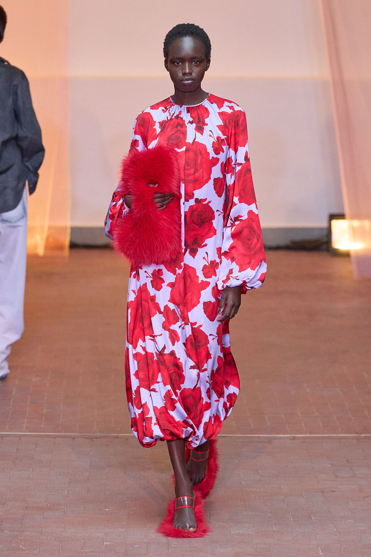 Model walking a runway in a white dress with large red rose print, holding a red fluffy clutch trailered by her side.
