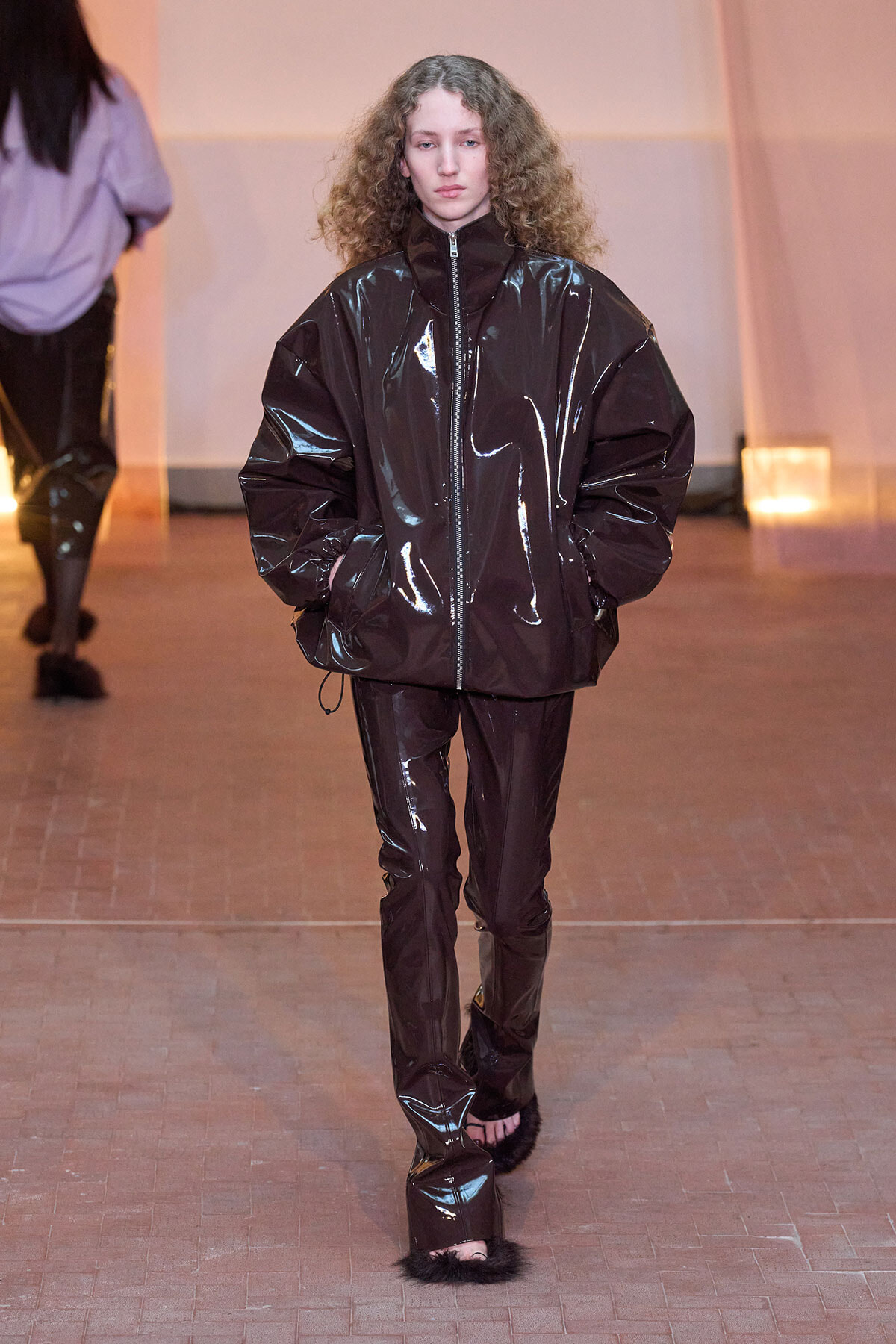 Model walks a runway in a glossy, oversized black zip-front jacket with matching pants and fur-trimmed footwear, under warm stage lighting.